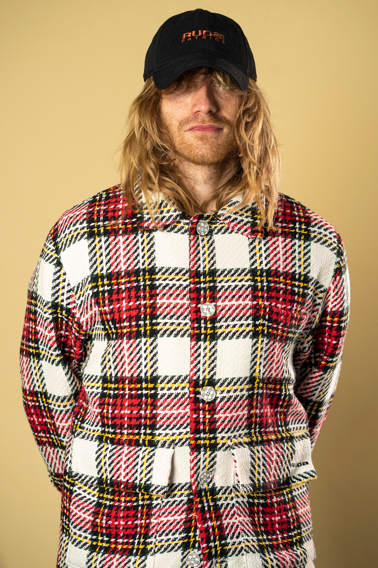 A person with long blond hair, possibly Ruda Patrice, is wearing a black hat and a Check Workers Jacket from Ruda Patrice. They are standing against a plain beige background.