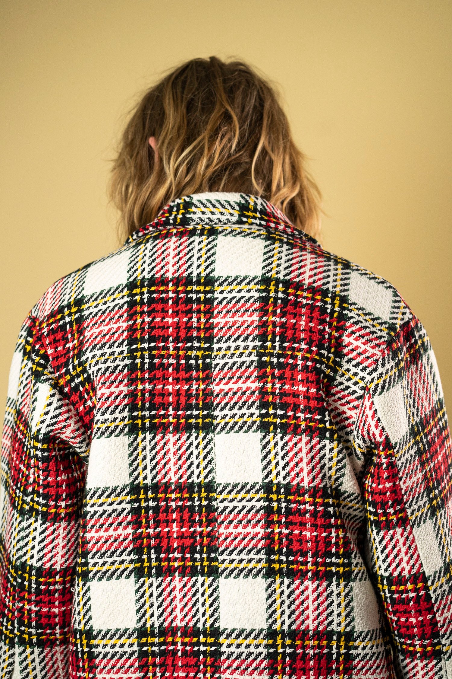 Rearr View: A person with wavy, shoulder-length hair is seen from the back, wearing a vibrant Ruda Patrice Check Workers Jacket. The jacket showcases a red, white, black, and yellow tartan pattern. The background is a solid warm beige color, and the person's head is turned slightly to the left.