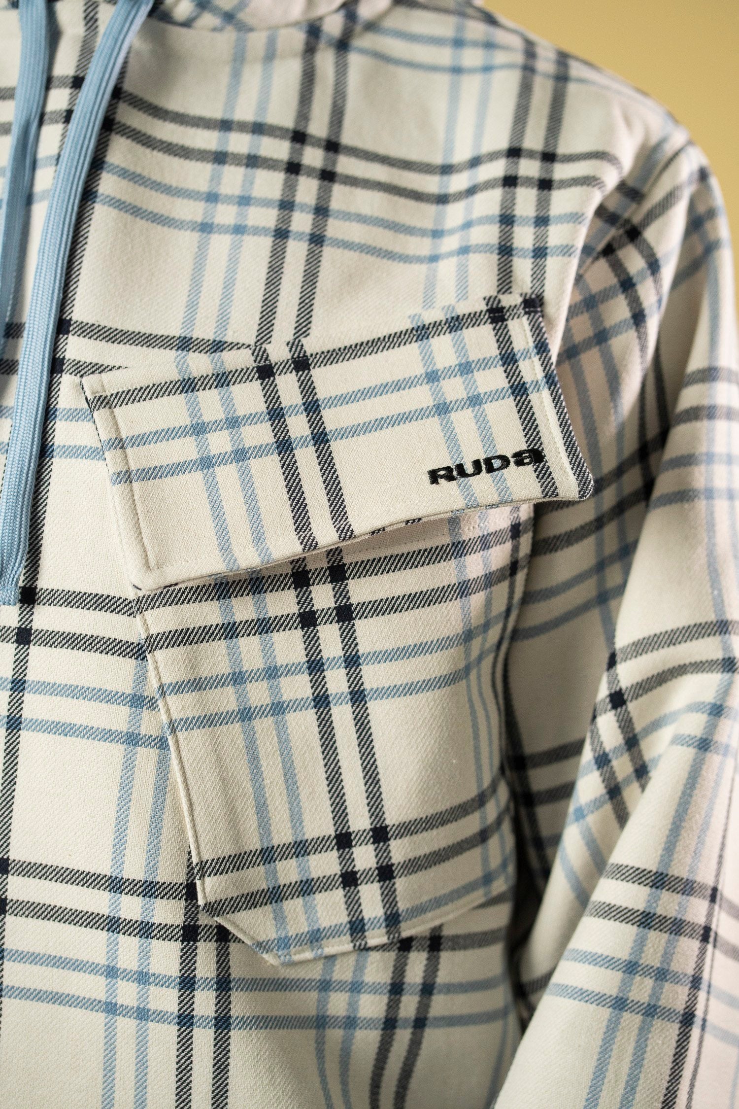 Close-up of a white and light blue plaid Blue Check Dead Stock Cotton Anorak featuring a front pocket on the chest. The pocket flap has the text "Ruda Patrice" printed on it in black letters, and there is a blue zipper extending from the collar.