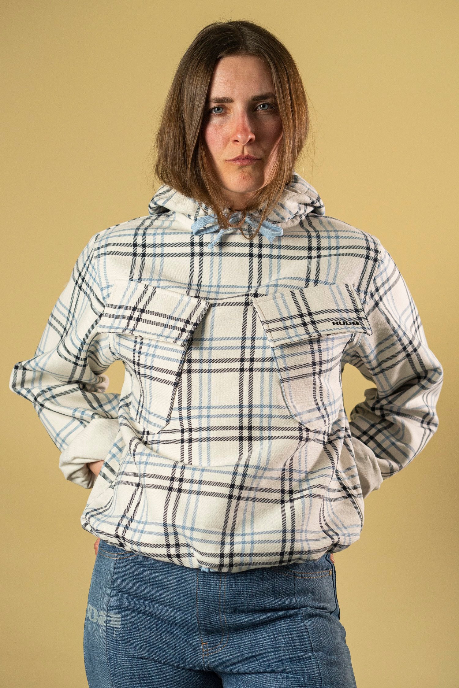 A person with long hair is standing against a beige background, wearing the stylish Ruda Patrice Blue Check Dead Stock Cotton Anorak, characterized by its blue and white checkered patterns. The anorak features two front pockets and a hood. They have their hands in the pockets and are looking directly at the camera.