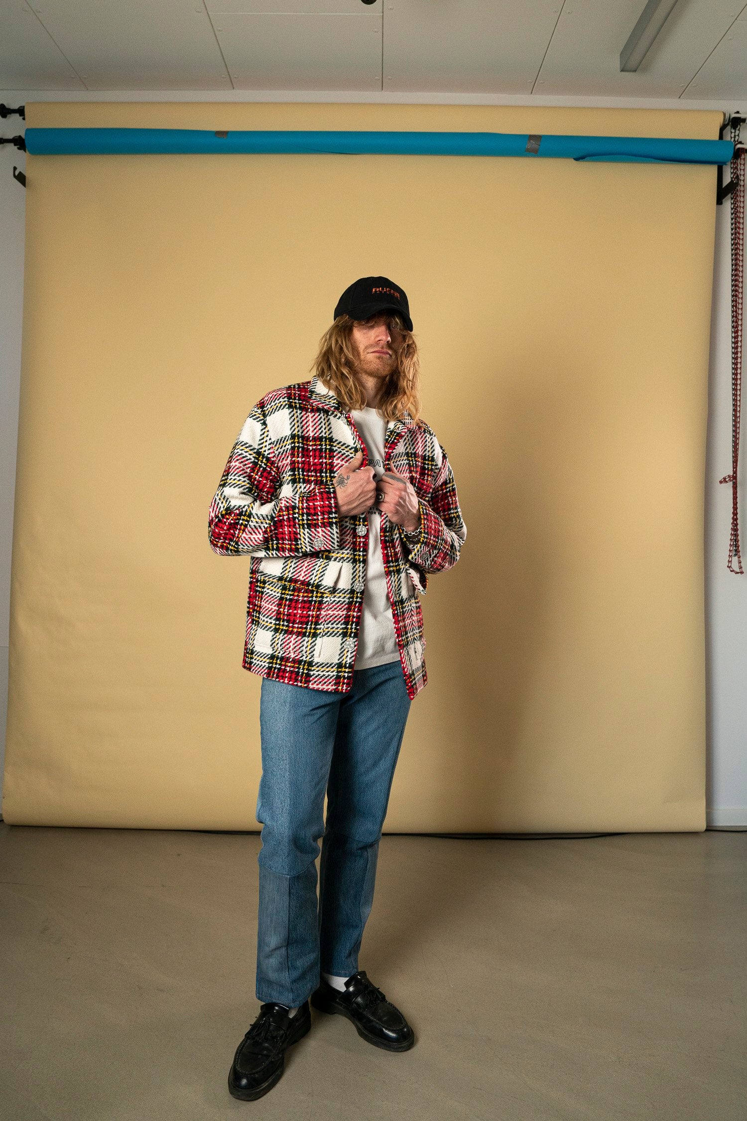 A person with long, blonde hair stands in front of a beige backdrop. They are wearing a black cap, the Check Workers Jacket by Ruda Patrice, a light grey shirt, blue jeans, and black sneakers. The individual is looking slightly downwards, holding the collar of their jacket with both hands—channeling a style reminiscent of Ruda Patrice.