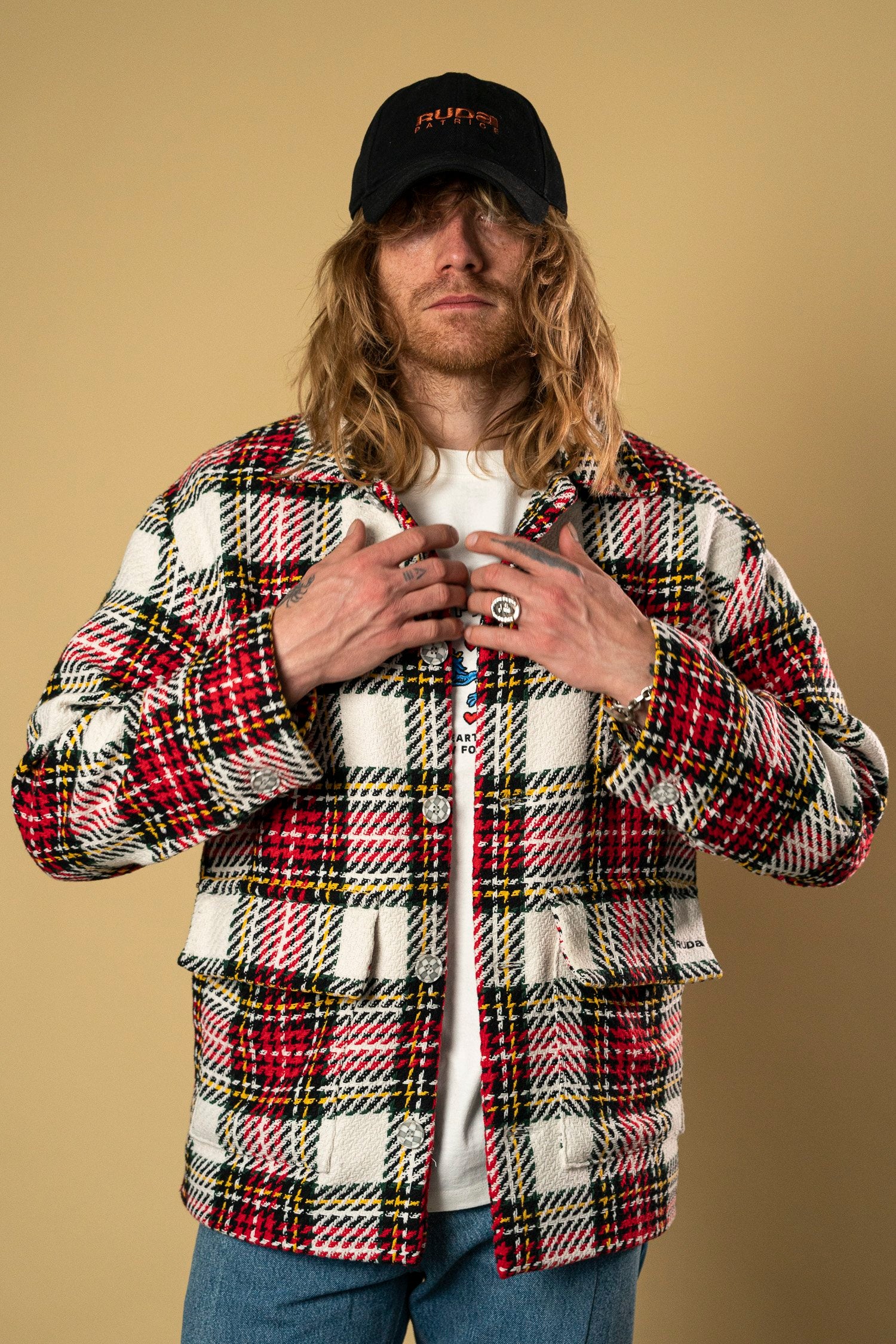 A person with long hair wearing a black baseball cap, a white t-shirt, and the Ruda Patrice Check Workers Jacket, featuring red, black, and yellow stripes, stands against a plain beige background. They have multiple rings on their fingers and are holding the edges of the jacket with an undeniable air of Ruda Patrice flair.