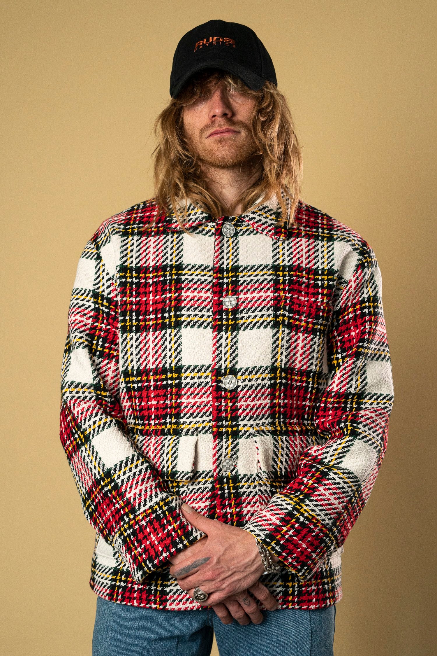 A person with long hair wearing a black cap and a Check Workers Jacket by Ruda Patrice, featuring a red, white, and black plaid design, stands against a plain beige background. They have their hands clasped in front of them and are also dressed in blue jeans.