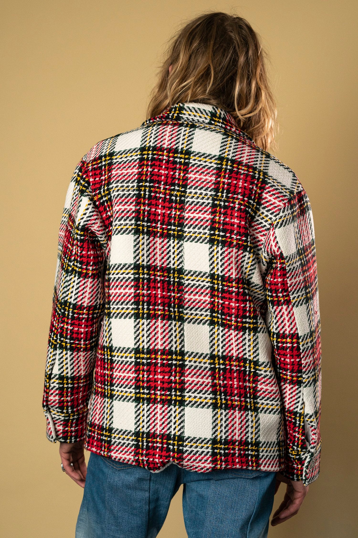 A person with long hair is wearing the Check Workers Jacket by Ruda Patrice, featuring a red, white, yellow, and black plaid pattern. The person is facing away from the camera, showcasing the back of the jacket against a plain beige background.