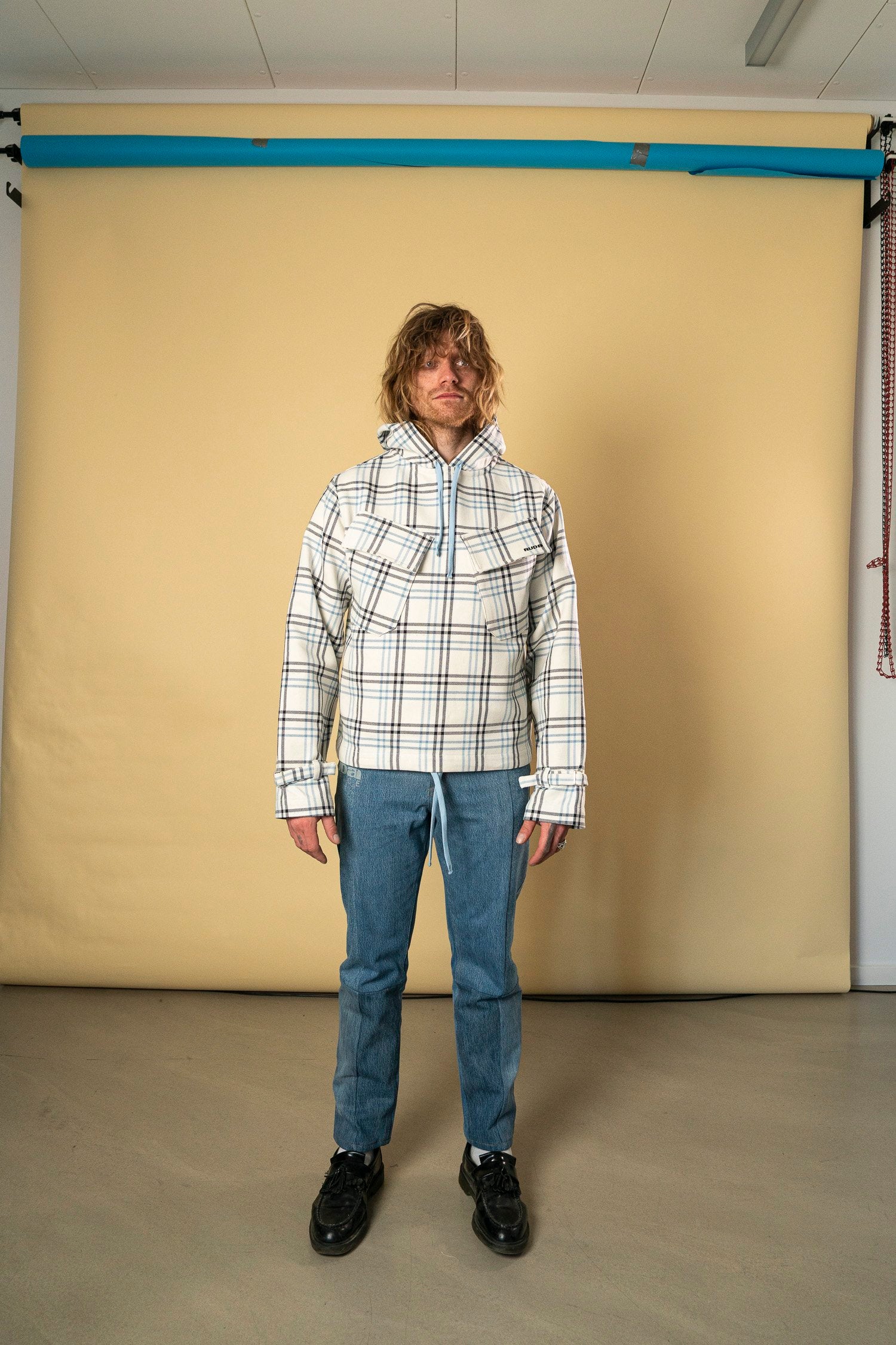 A person with long blond hair stands in front of a beige backdrop. They are wearing Ruda Patrice's Blue Check Dead Stock Cotton Anorak, blue jeans, and black shoes. With a relaxed posture, they embody the laid-back style often showcased by Ruda Patrice in an indoor studio setting.