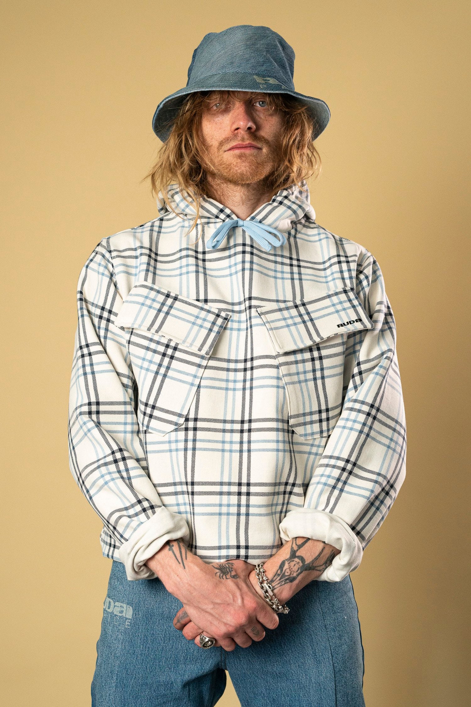 A person with long hair wearing a Blue Check Dead Stock Cotton Anorak, denim bucket hat, and denim jeans stands against a beige background. Their hands are clasped together, displaying several tattoos and bracelets. The Ruda Patrice anorak has front pockets, and the individual exudes a confident expression.