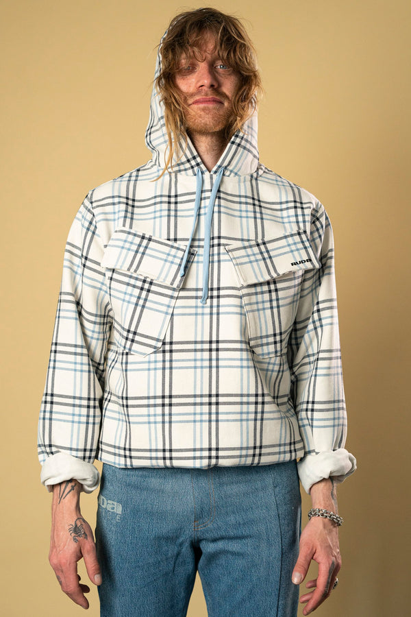 A person with long, tousled hair and a touch of Ruda Patrice style wears the Blue Check Dead Stock Cotton Anorak—a hooded pullover with a white and blue plaid pattern, half-zip, and large chest pocket. They have tattoos on their hands and wrists that complement their blue jeans. The background is a plain beige color.