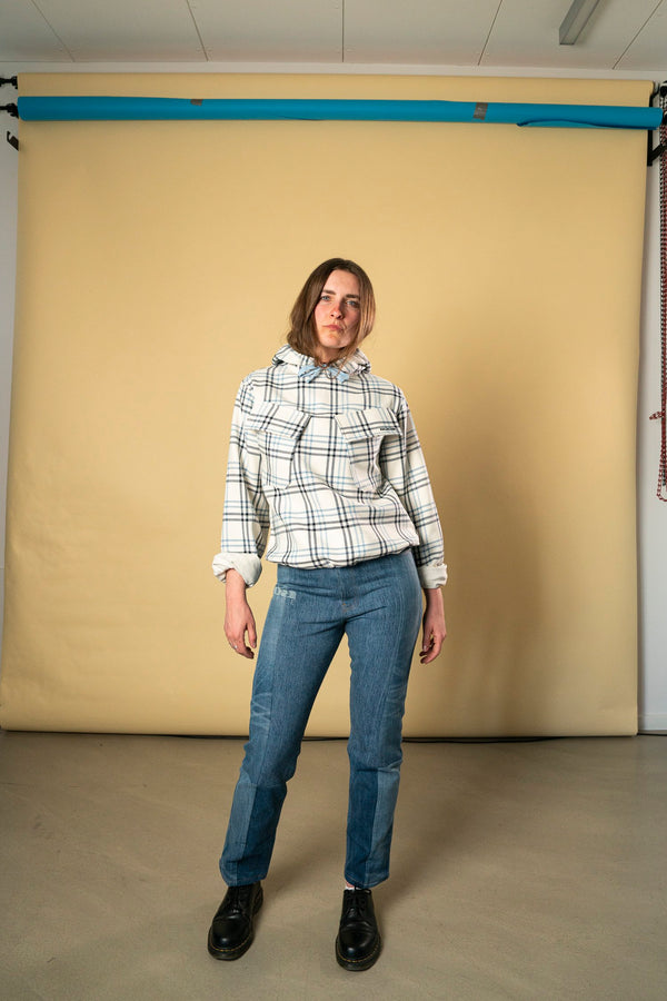 A person stands indoors in front of a beige backdrop. They are wearing the Blue Check Dead Stock Cotton Anorak from Ruda Patrice, paired with blue jeans and black boots. Their hands are relaxed at their sides, and their head is slightly tilted in a pose reminiscent of Ruda Patrice's signature style.