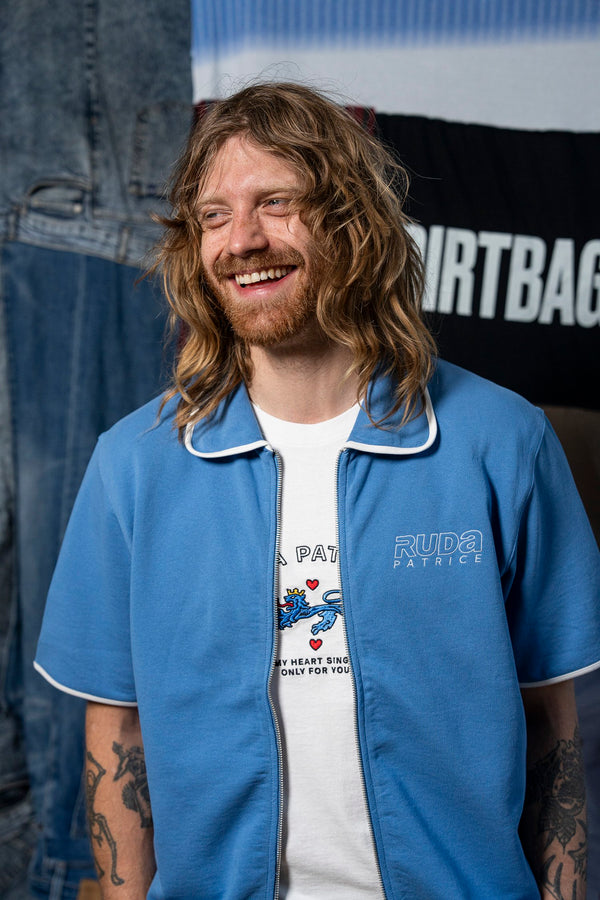 A man with long, wavy blond hair and a beard smiles while wearing the Maguire Shirt - Blue from Ruda Patrice over a white t-shirt with a graphic design. Tattoos are visible on his arms. Behind him, denim fabric and a banner with partially visible text reading "Ruda Patrice" are seen.