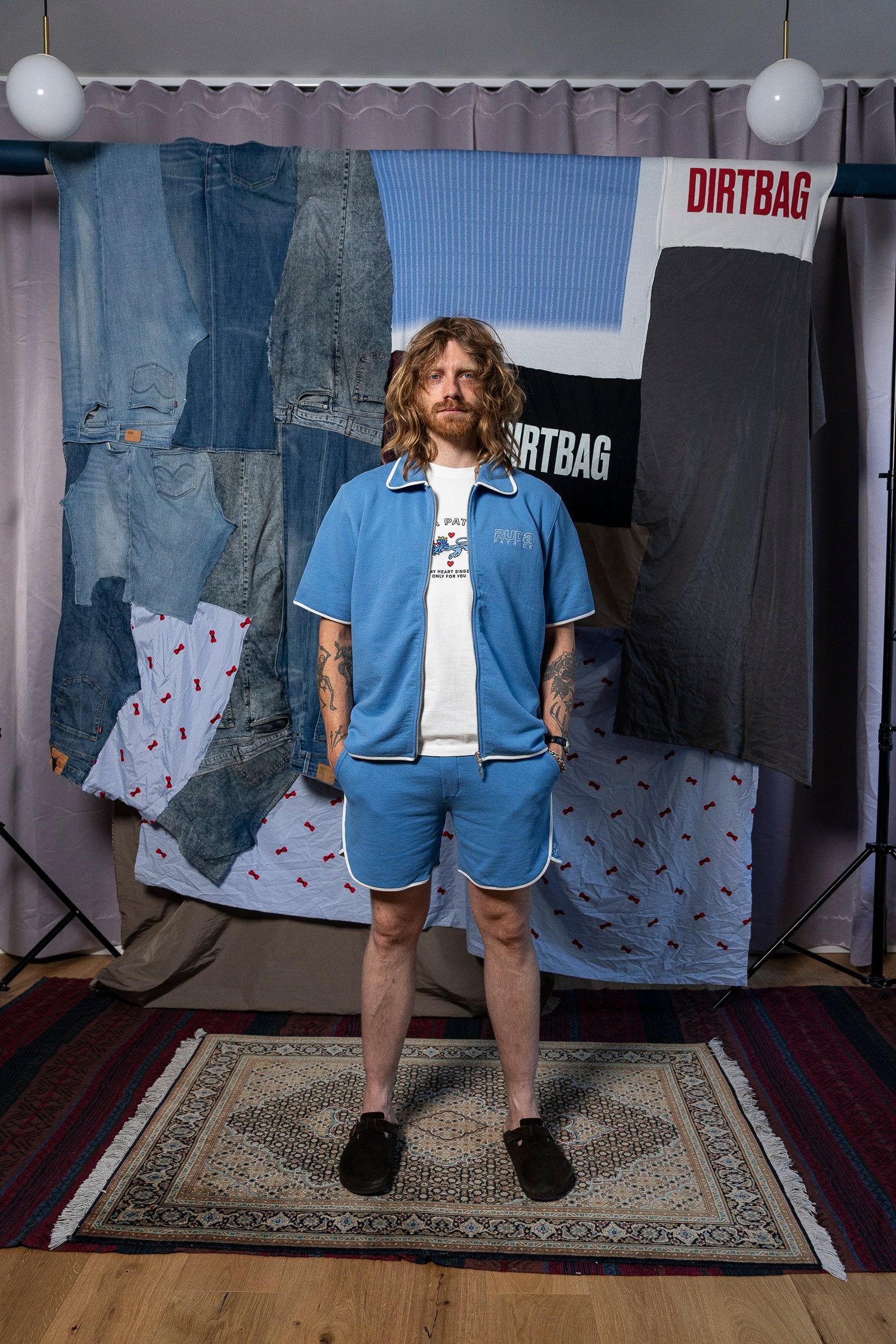 A person with long, wavy hair and tattoos stands in front of a multi-fabric backdrop, wearing the Maguire Short - Blue by Ruda Patrice. They have on a light blue shirt and matching shorts with a white T-shirt underneath. The backdrop includes denim and patterned fabrics, prominently displaying the word "DIRTBAG," creating a stylish scene that feels like it could belong to Ruda Patrice's latest collection.