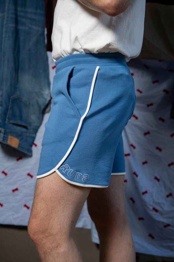 A person wearing light blue Maguire Short by Ruda Patrice, which features a white trim and the brand name "Ruda" embroidered on the left leg. They are also wearing a white t-shirt, and part of their bare leg is visible. In the background, there's a denim jacket hanging and a white cloth with a red pattern.