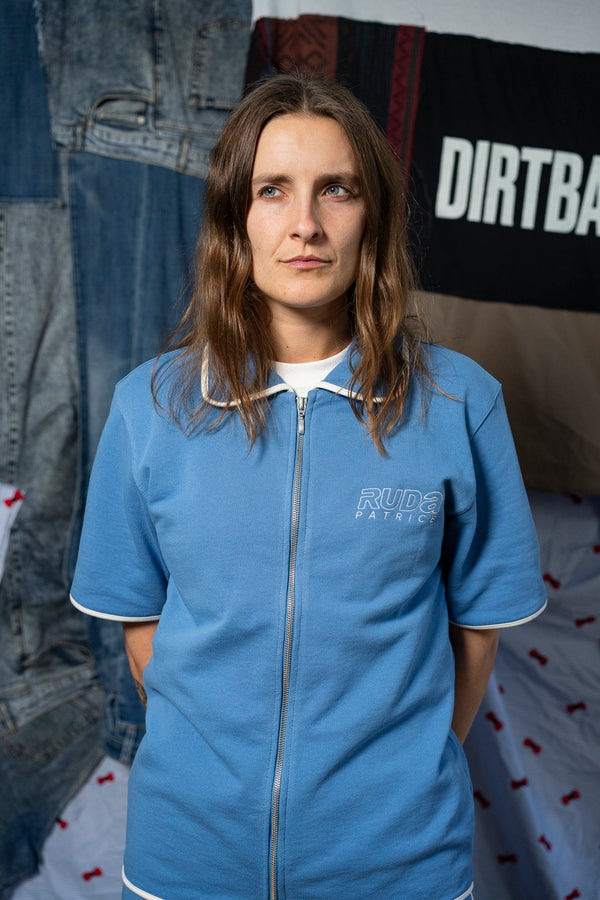 A person with long, brown hair stands facing forward, wearing a Maguire Shirt - Blue by Ruda Patrice. The background features various fabrics, including denim and a black and white fabric with the text "DIRTBA" partially visible.