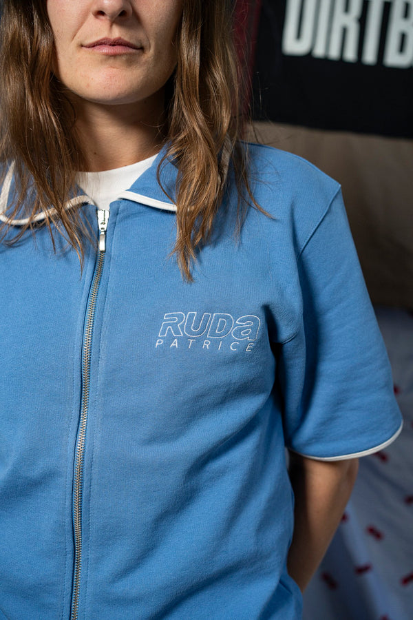 A person with long hair is wearing the Maguire Shirt in blue, featuring white piping on the sleeves and collar. The shirt has "Ruda Patrice" embroidered on the chest. A portion of a partially visible banner with indistinct text is in the background.