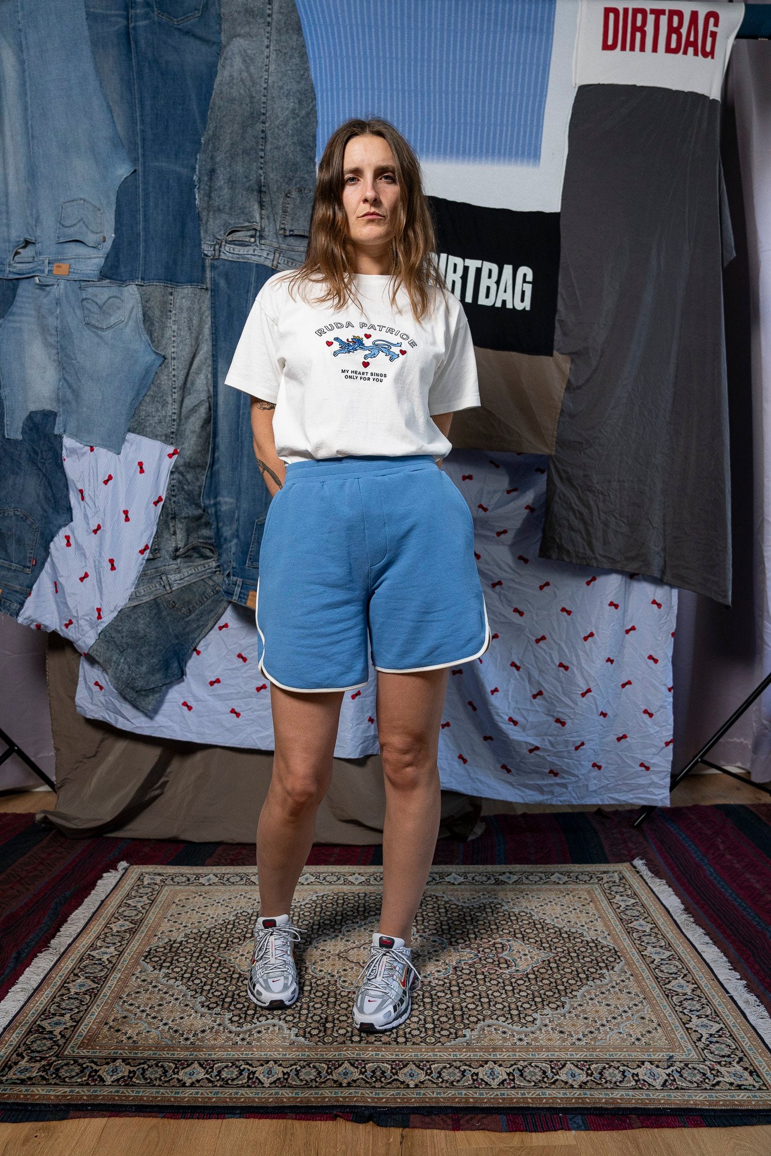 A woman stands on a patterned rug wearing a white graphic T-shirt, the Maguire Short in blue with white trim from Ruda Patrice, and white athletic shoes. Behind her is a backdrop of denim and fabric pieces, with two clear "DIRTBAG" logos visible. She has long light brown hair and a neutral expression, effortlessly embodying the Ruda Patrice style.