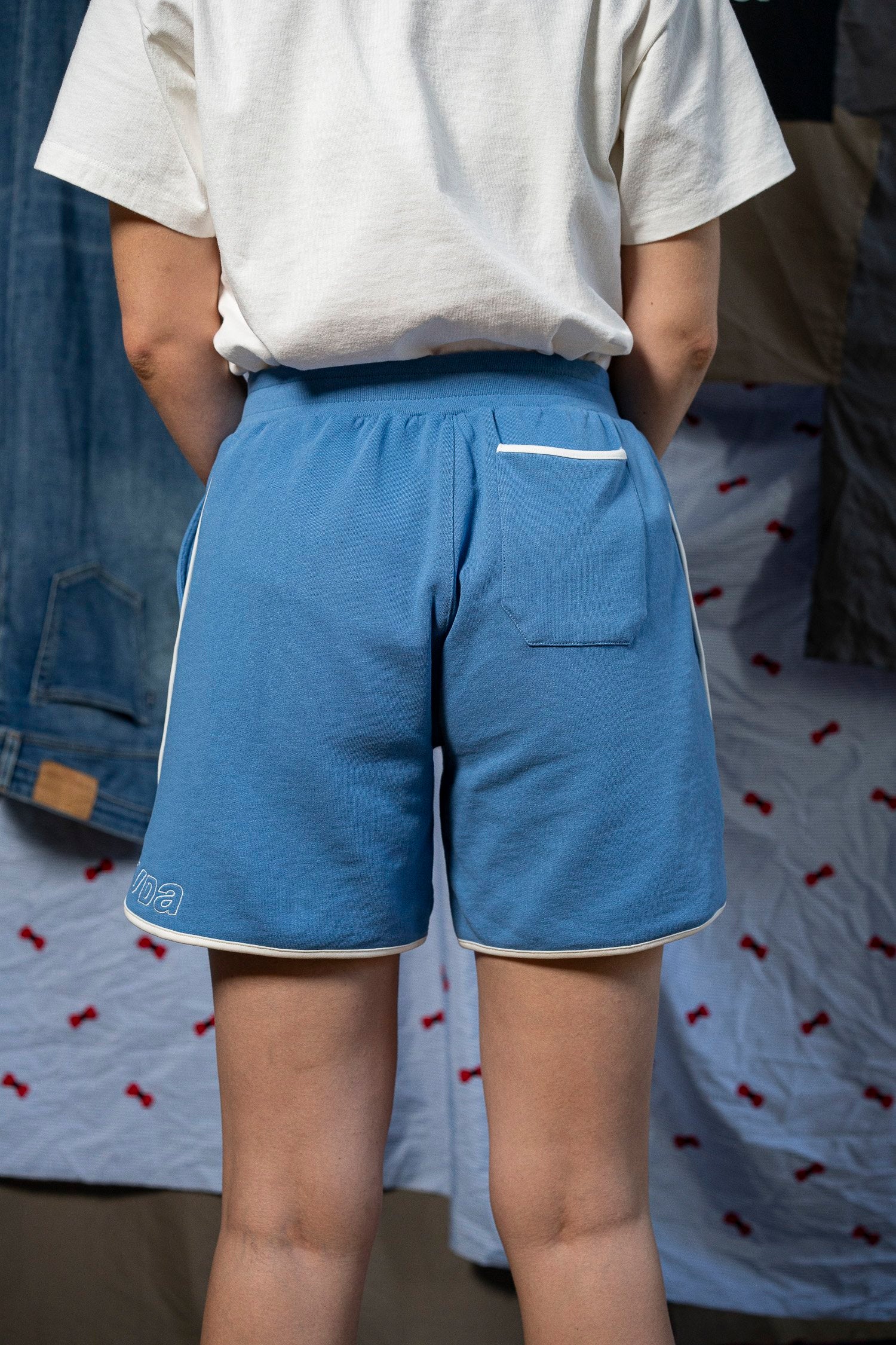 A person is seen from behind, wearing the Maguire Short - Blue by Ruda Patrice, featuring white trim and a small pocket on the back. The shorts have a Ruda Patrice logo on the left. The individual is also wearing a white t-shirt. In the background, there are various hanging fabrics.