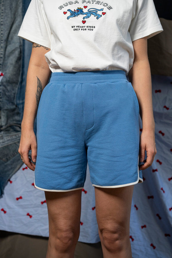 A person is wearing a white t-shirt featuring text and graphics, paired with the Maguire Short - Blue from Ruda Patrice, which have white trim. They have a tattoo on their right forearm. The background contains a mix of fabrics, including a denim jacket designed by Ruda Patrice and a cloth with red bow patterns.